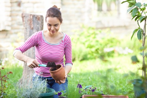 A woman with dark hair tied back, wearing a pink and white striped long-sleeved top, is engaged in gardening in a lush backyard garden. She is holding a terracotta pot filled with dark soil, preparing to plant or transplant flowers. The garden features a well-maintained lawn with vibrant green grass, bordered by flower beds with purple and green foliage. In the background, there are small shrubs, a tree trunk, and a brick wall with some out-of-focus greenery, suggesting a private outdoor space in Northolt. The weather is bright and clear, providing natural light that illuminates the scene. Gardening tools and plant pots are visible on the ground, indicating active gardening work. The scene reflects outdoor maintenance and gardening activities typical of residential gardens in the Northolt area, as part of professional gardening services provided by Gardening Services Northolt to enhance outdoor spaces.