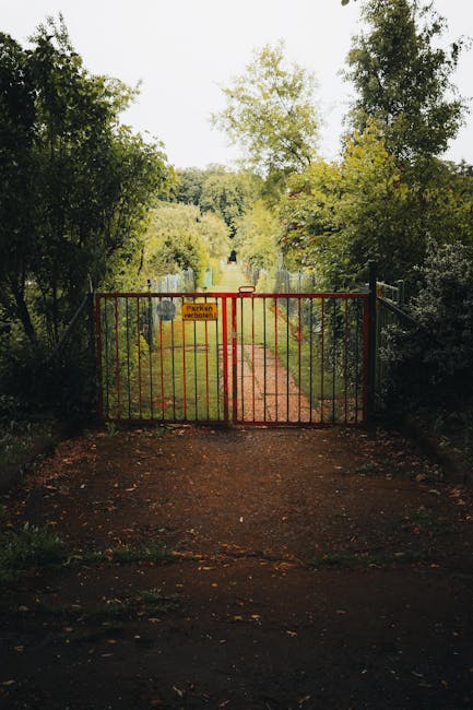 A small garden gate made of vertical wooden slats painted in a deep red color, with a diagonal support beam for stability, attached to a metal frame at the top. The gate is partially open, revealing l